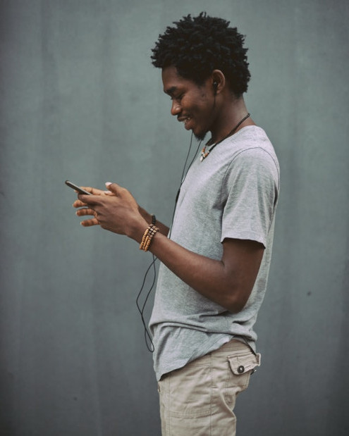 Seorang pria berdiri di dinding abu-abu memegang earphone dan teleponnya.